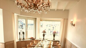 Una larga mesa de comedor preparada para una comida, con sillas, una araña y ventanas con cortinas.