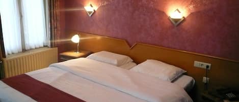 Una habitación de hotel con una cama grande, dos lámparas de noche, un ventanal con cortinas y un radiador.