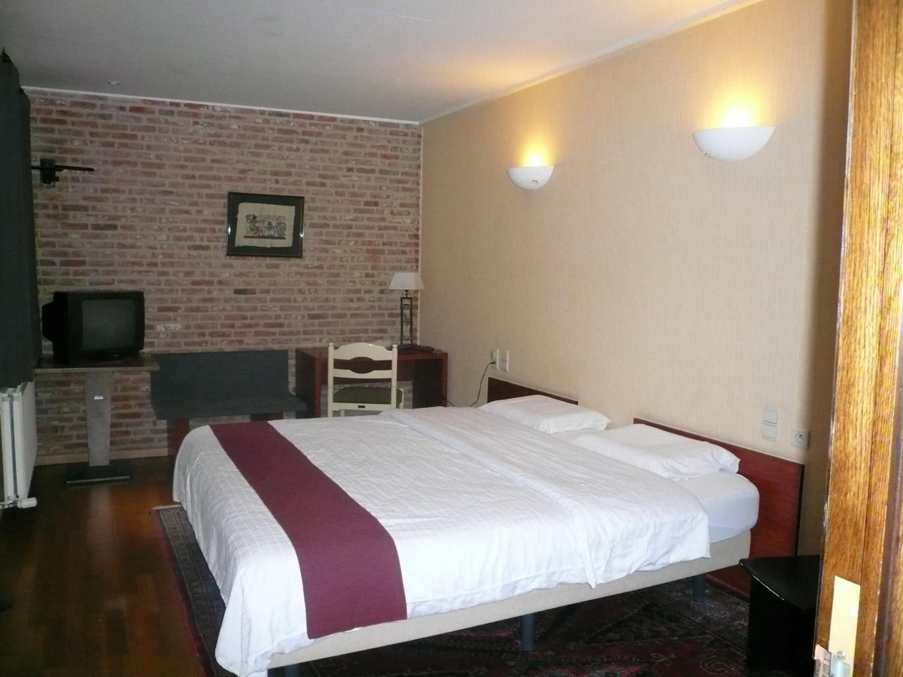 Habitación de hotel con pared de ladrillo, cama con ropa de cama blanca y un detalle rojo, un televisor sobre un mueble y dos luces fijadas a la pared.
