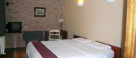 Habitación de hotel con pared de ladrillo, cama con ropa de cama blanca y un detalle rojo, un televisor sobre un mueble y dos luces fijadas a la pared.