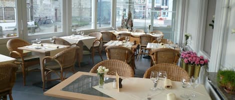 A restaurant interior with multiple tables and wicker chairs, large windows offering a view of the street, and a variety of table settings.