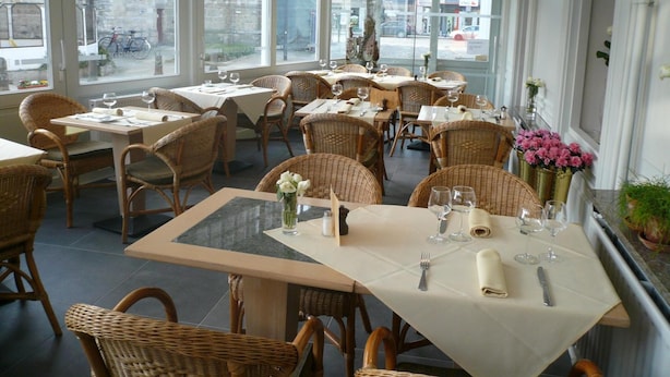 Un interior de restaurante con varias mesas y sillas de mimbre, grandes ventanales que ofrecen vistas a la calle y diferentes tipos de mesas dispuestas.