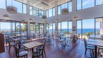 A spacious indoor dining area with large windows offering ocean views, wooden flooring, and modern pendant lighting.