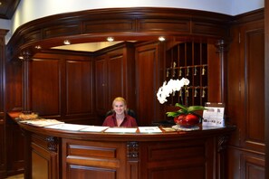 Een receptiebalie met een vrouw erachter, omringd door houten lambrisering en een glazen vitrine met sleutels.