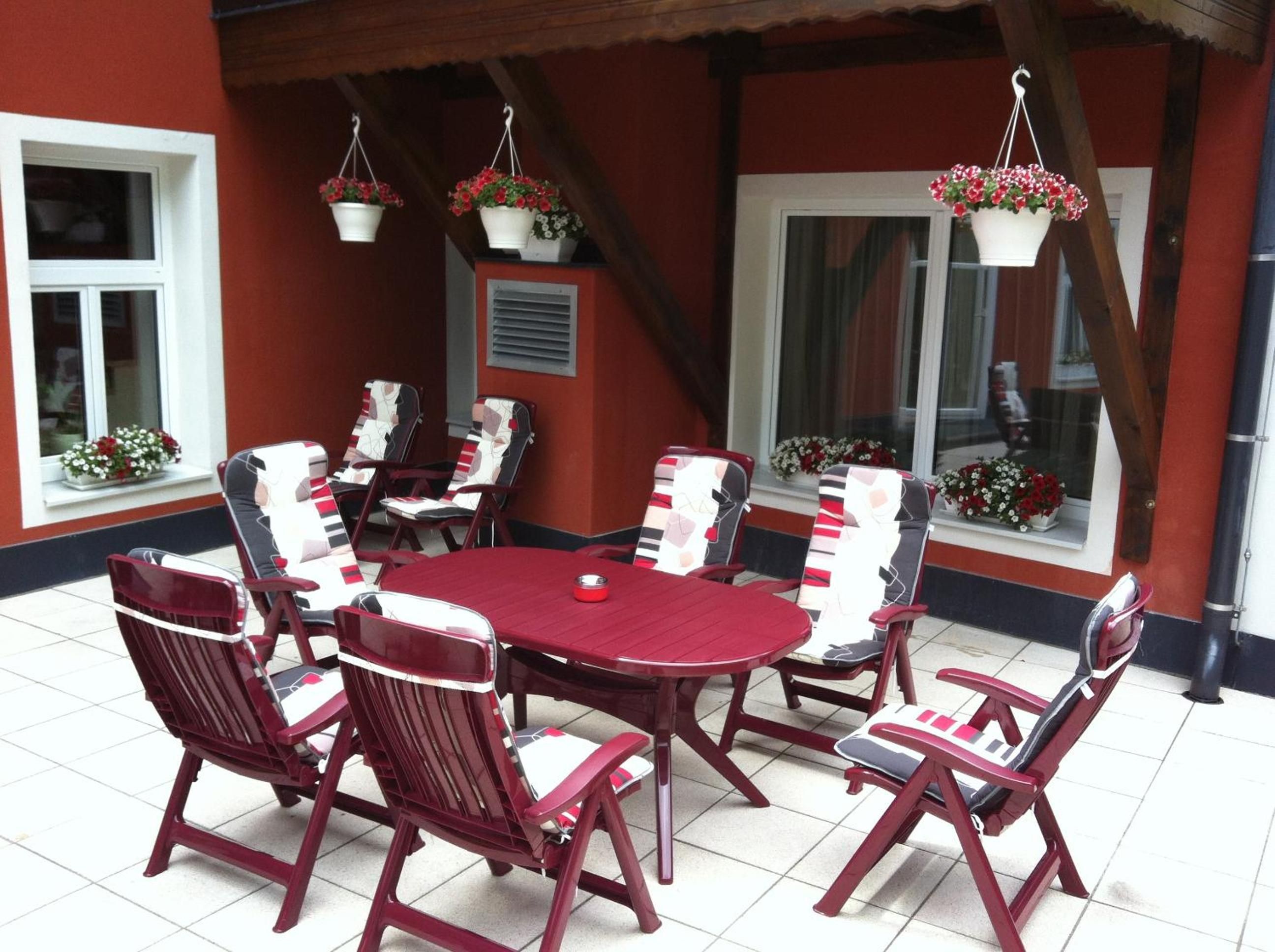 Un patio con tejas rojas, una mesa redonda y sillas a juego, rodeado de cestas colgantes con flores.