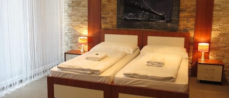 A double bed with a wooden frame and white bedding, two bedside tables with lamps, a cityscape painting on the wall, and a window with blinds.