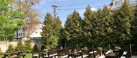 An outdoor seating area with wooden tables and benches, surrounded by potted plants and trees.