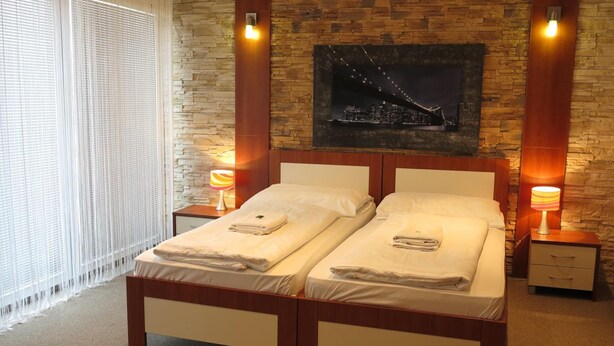 Una cama doble con estructura de madera y ropa de cama blanca, dos mesitas de noche con lámparas, un cuadro de paisaje urbano en la pared y una ventana con persianas.