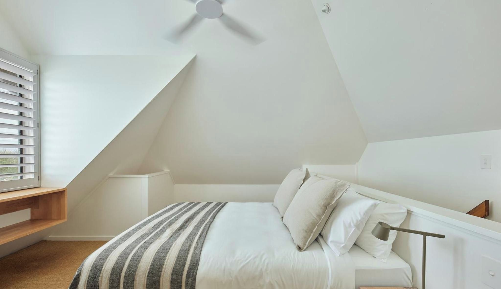 Un dormitorio con techo inclinado, una cama con ropa de cama blanca, una mesita de noche y una ventana con persianas.