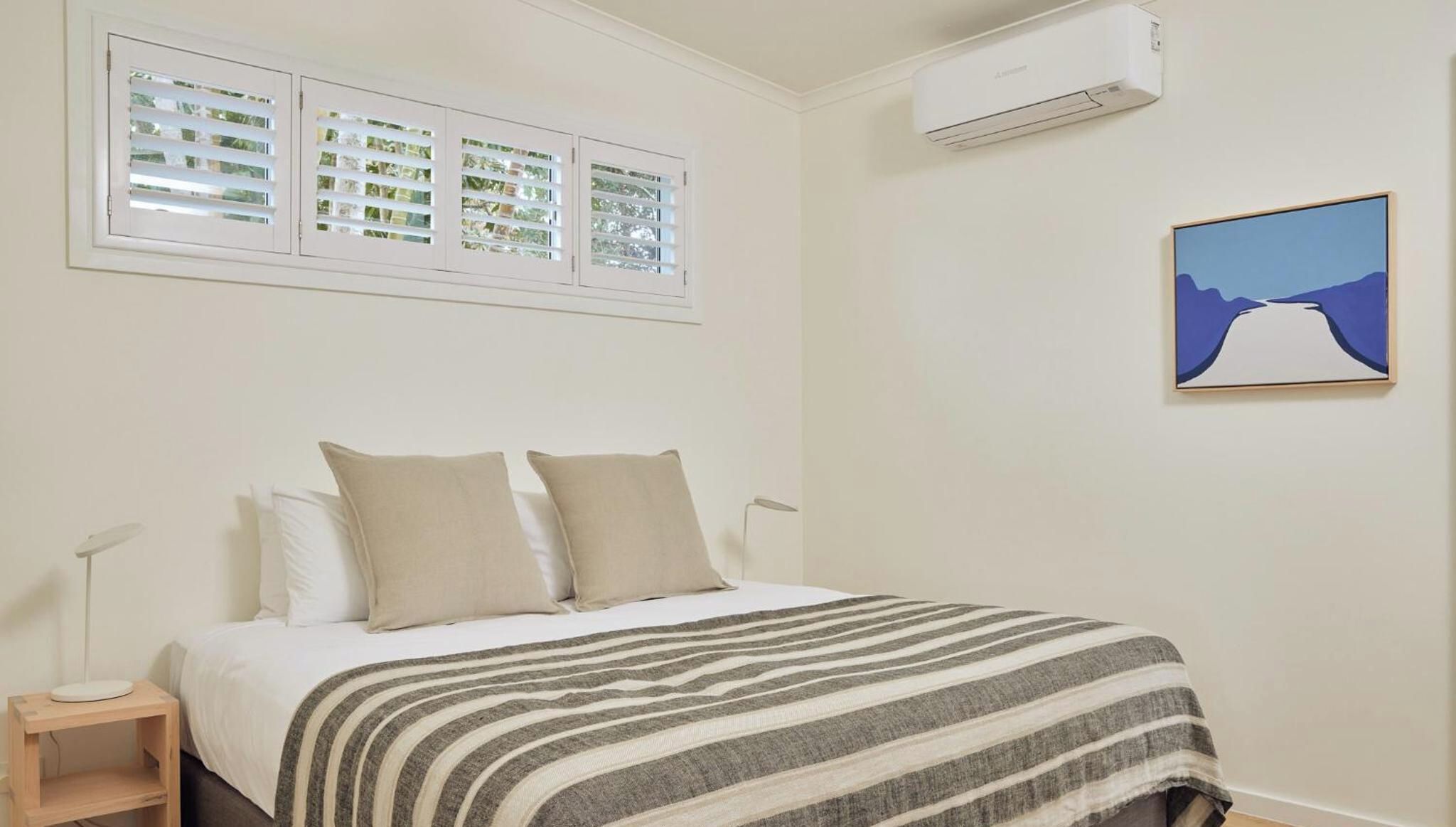 A bedroom with a bed, two pillows, a striped blanket, a window with blinds, an air conditioner, and a framed picture on the wall.