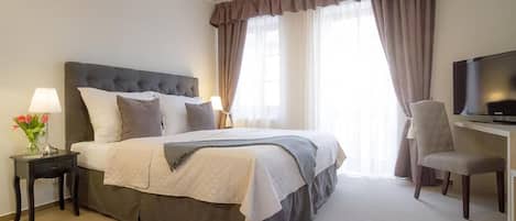 Quarto de hotel com uma cama grande, uma mesa com cadeira, televisão e mesa de cabeceira com abajur e flores.