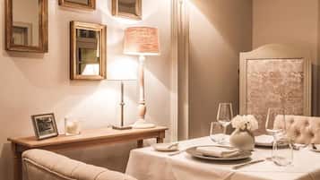 A dining area with a table set for two, a lamp, a mirror, and a framed picture on a wooden side table.