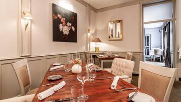 A dining room with a wooden table set for six, surrounded by chairs, a vase with flowers, and a painting on the wall.