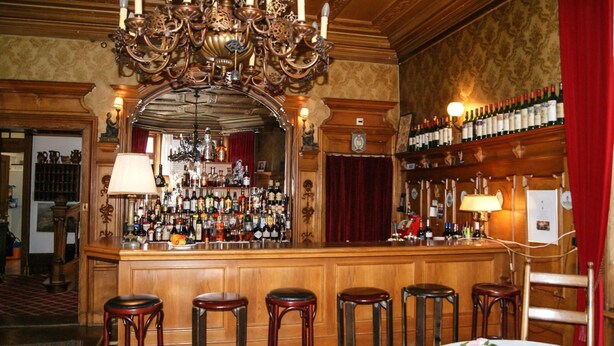 A bar area with a wooden counter, multiple stools, a chandelier, and a wine rack.