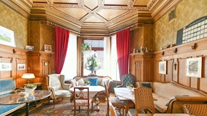 A traditional room with a high ceiling, ornate wood paneling, a large window with red curtains, and a variety of seating options including a sofa, chairs, and a table.