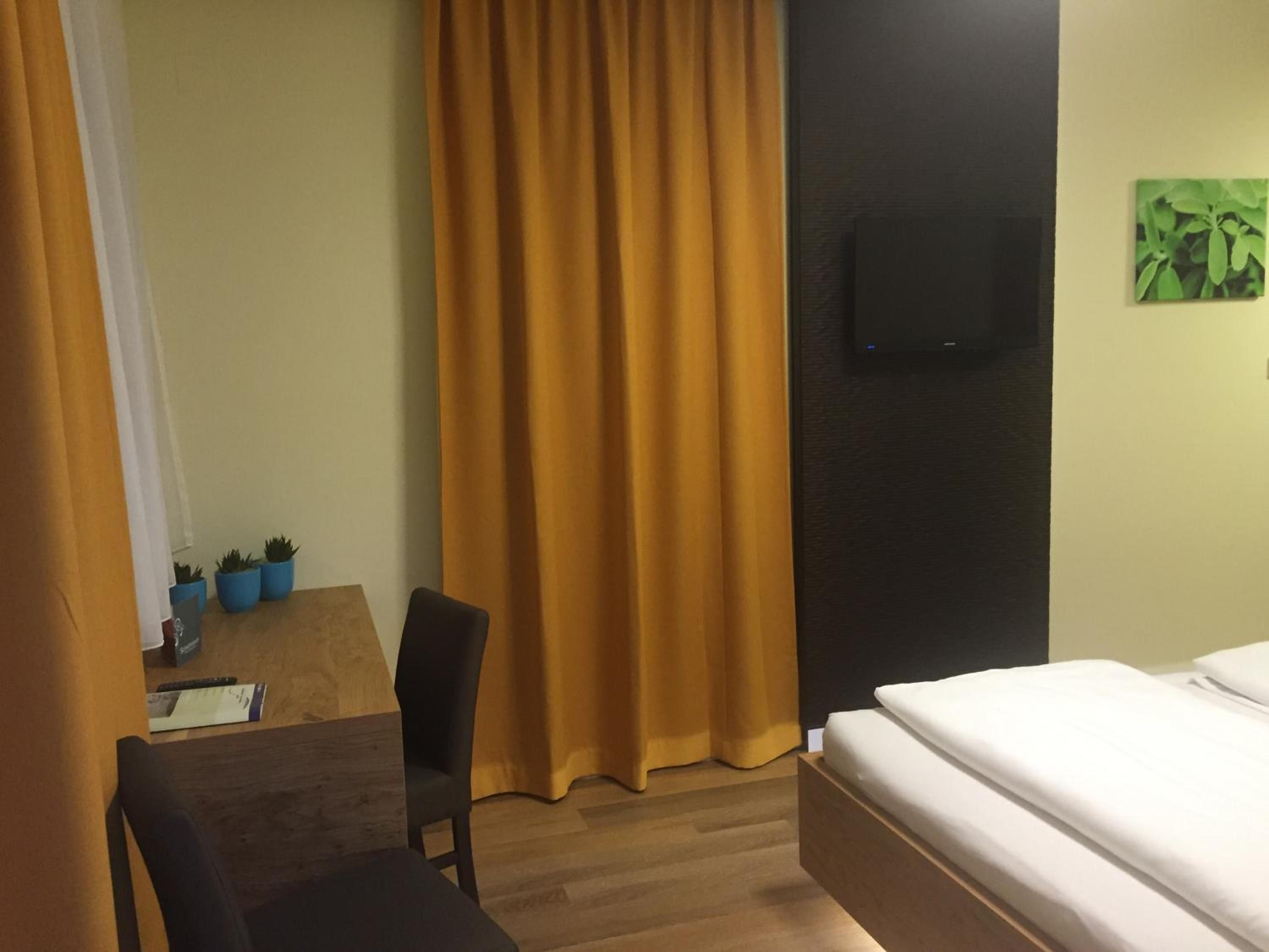 A hotel room with a bed, a desk with a chair, a television mounted on the wall, and a window with curtains.