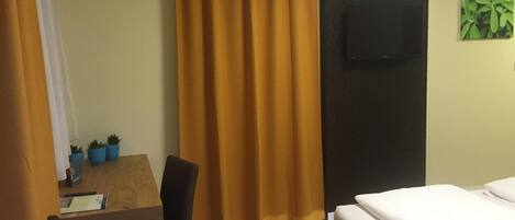A hotel room with a bed, a desk with a chair, a television mounted on the wall, and a window with curtains.