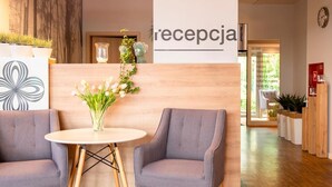 Uma área de recepção com balcão de madeira, duas poltronas cinzas, uma mesa redonda com um vaso de flores e uma placa na parede com a inscrição 'reception'.