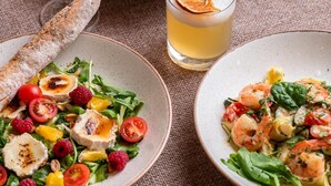 Uma mesa posta com diversos pratos, incluindo uma tigela de sopa, um prato de salada, um prato com camarões e vegetais, um copo de bebida transparente e um pão.