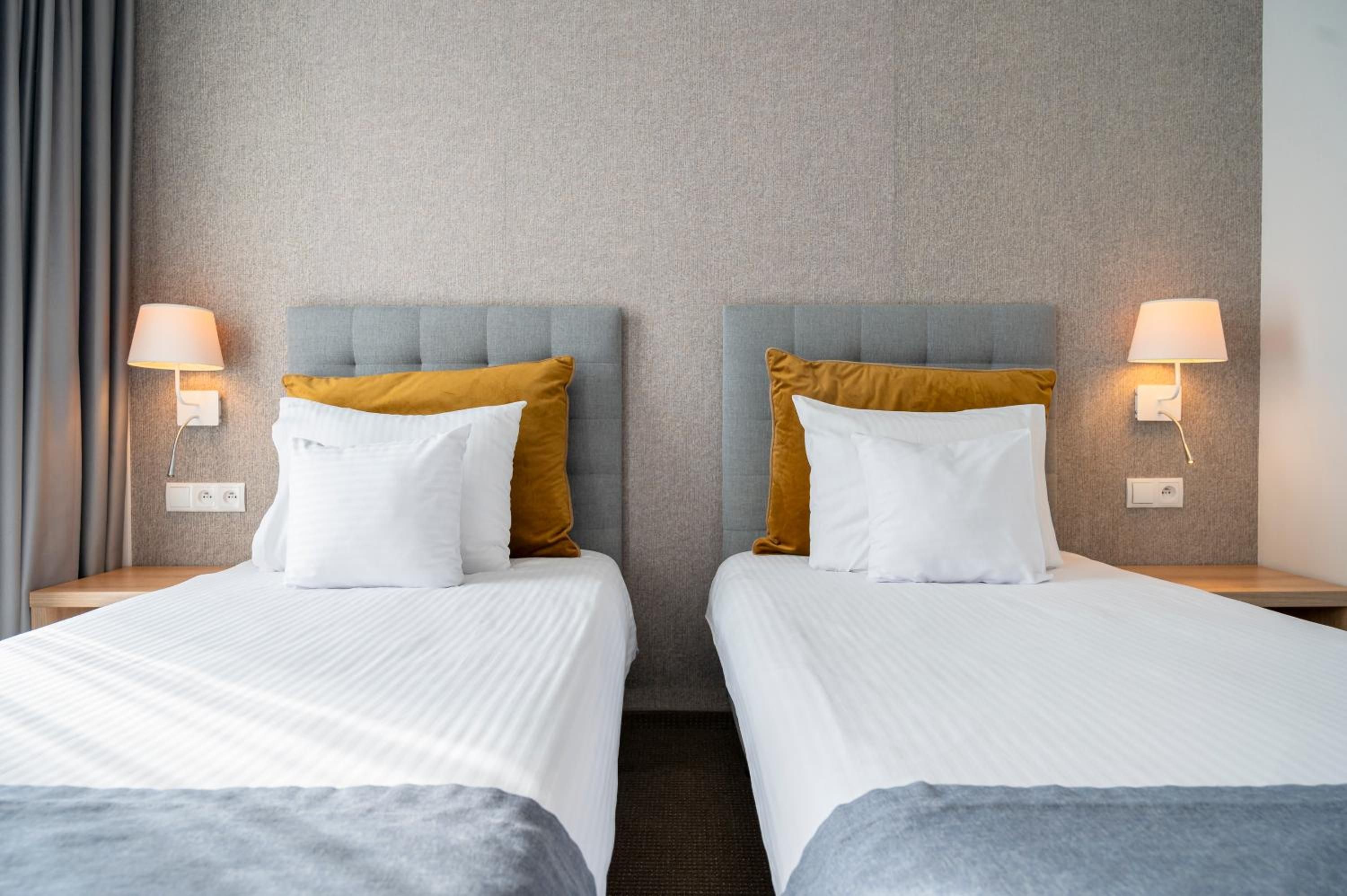 A hotel room with two beds, each with a white bedspread and a mustard-colored pillow. There are wall-mounted lamps on either side of the beds.