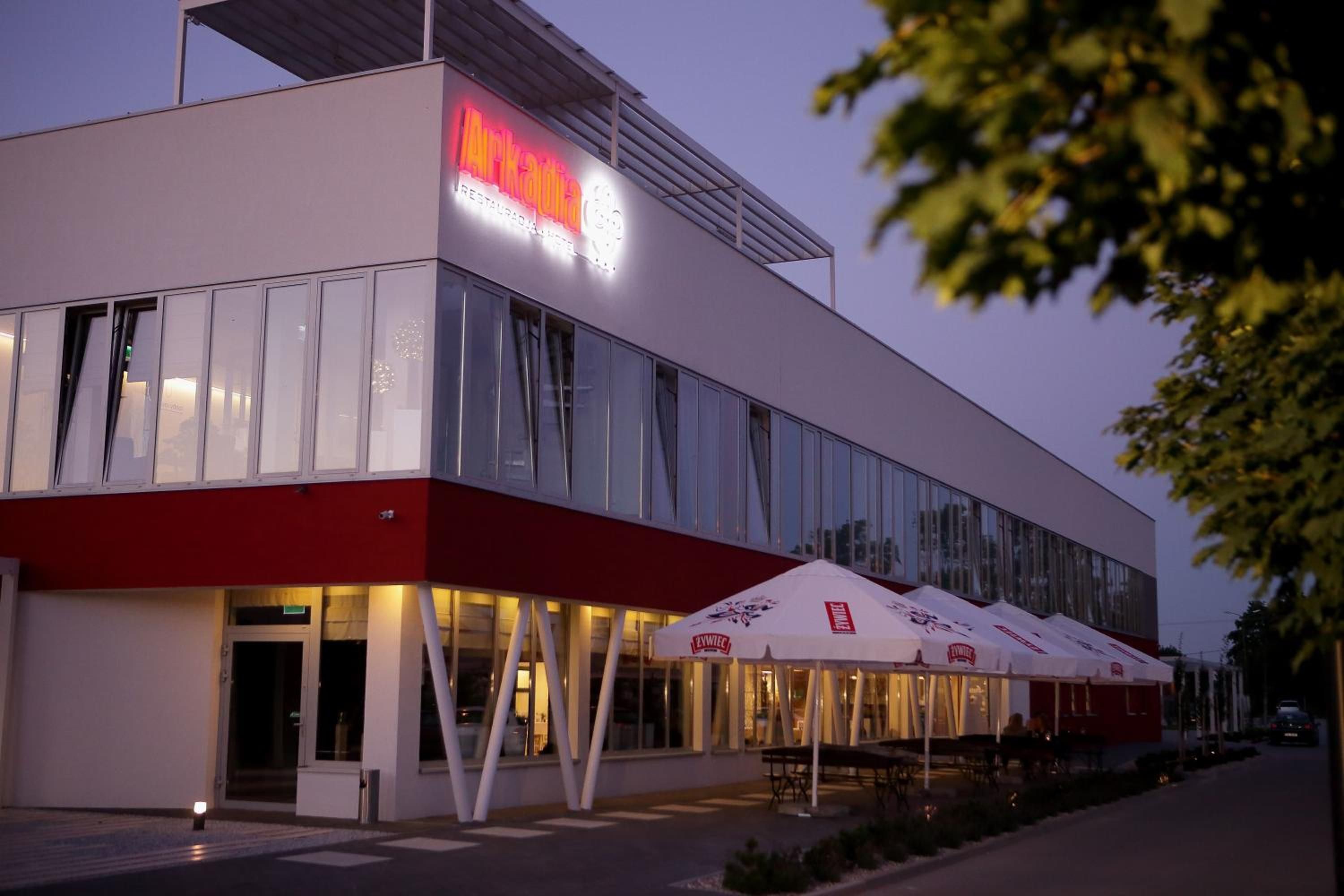 A modern building with a restaurant named 'Arkadia' on the ground floor, featuring large windows and outdoor seating with umbrellas.