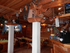 Een rustiek houten interieur met houten banken, een bar met glazen en flessen, en een hangklok.