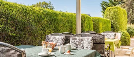 An outdoor patio with a table set for tea, surrounded by chairs with patterned cushions, under a clear sky.