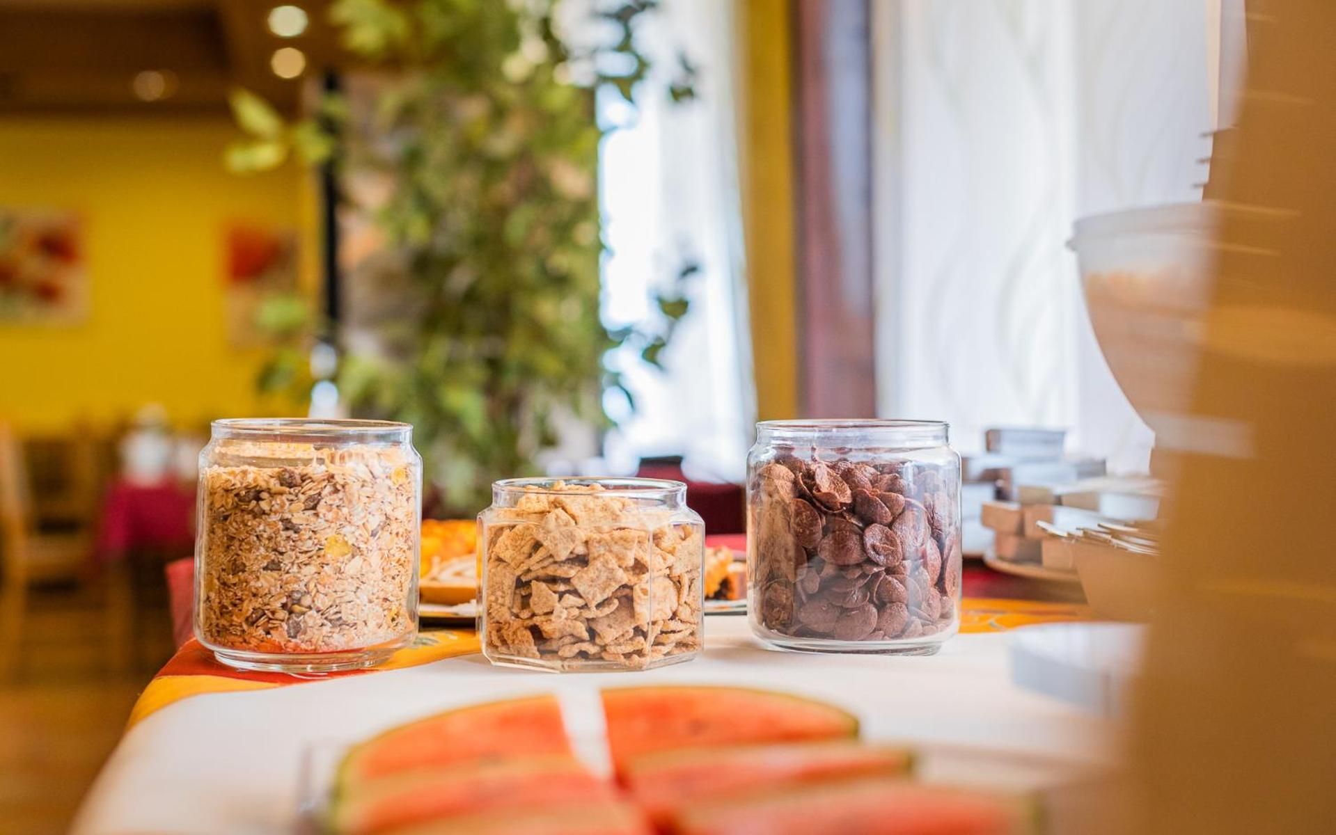 Three glass jars containing different types of cereal on a table with a slice of watermelon.