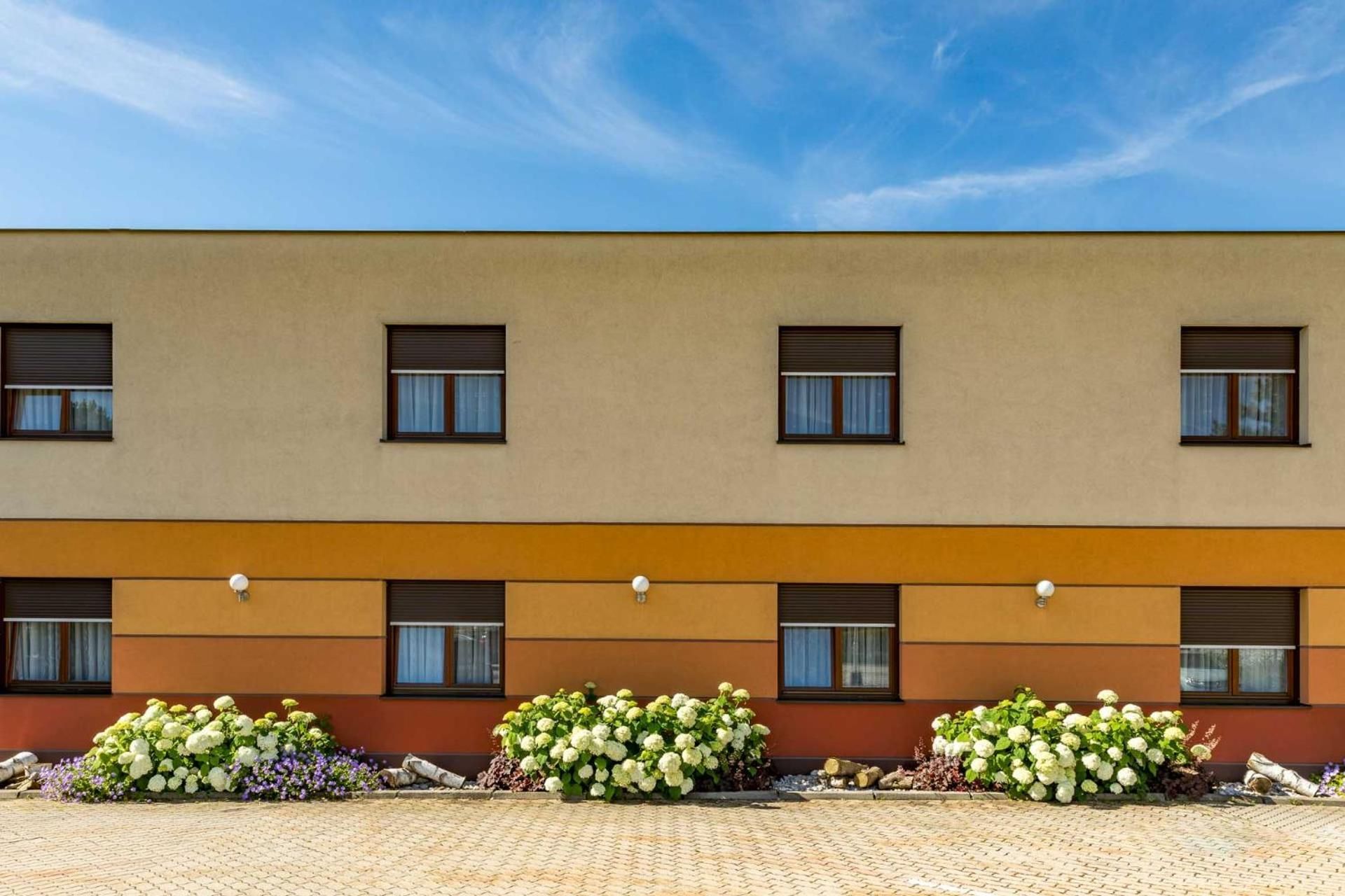 A building with a flat facade, multiple windows, and a paved ground with flower beds.