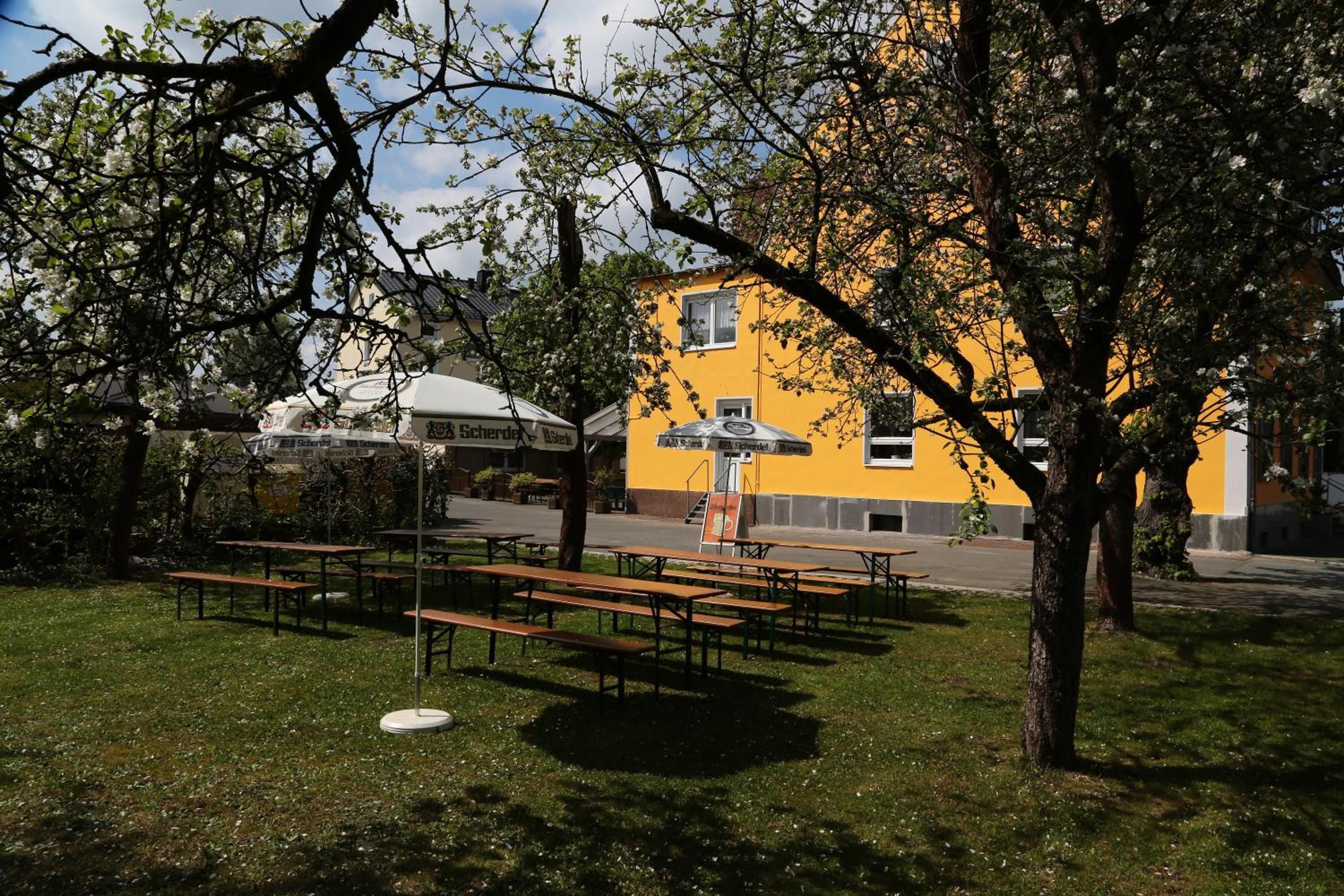 Un área de estar al aire libre con mesas de picnic y bancos, rodeada de árboles y un edificio amarillo con un letrero.