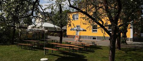 Un área de estar al aire libre con mesas de picnic y bancos, rodeada de árboles y un edificio amarillo con un letrero.