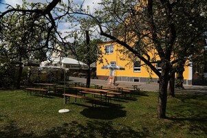 Un área de estar al aire libre con mesas de picnic y bancos, rodeada de árboles y un edificio amarillo con un letrero.