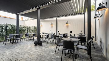 An outdoor patio area with tables and chairs, a pergola structure, and wall-mounted lighting.