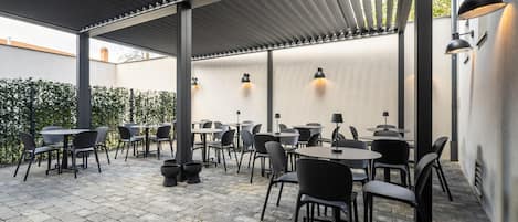 An outdoor patio area with tables and chairs, a pergola structure, and wall-mounted lighting.