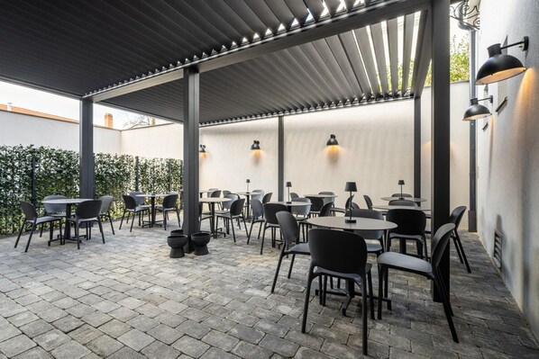 An outdoor patio area with tables and chairs, a pergola structure, and wall-mounted lighting.