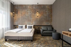 A modern hotel room with a large bed, a grey sofa, and a patterned carpet.