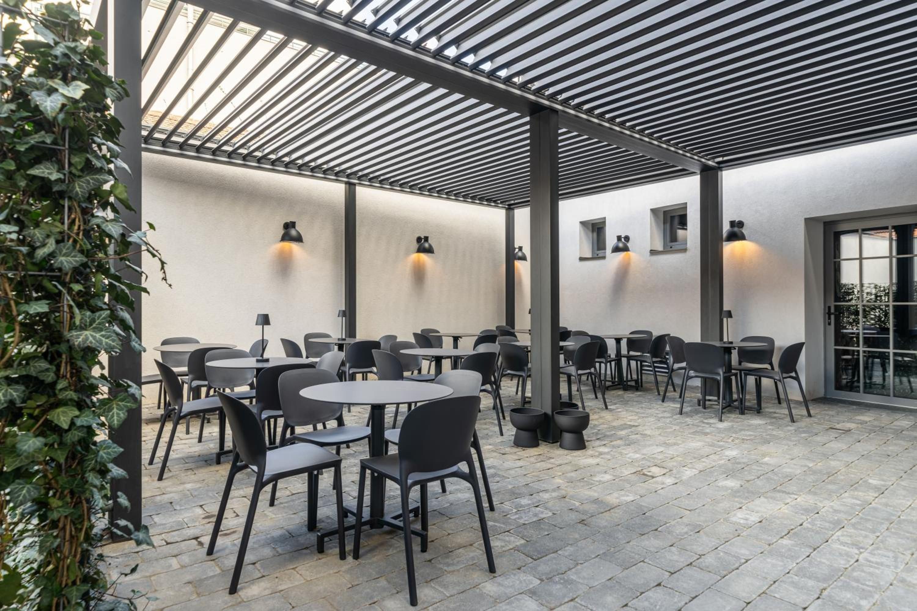 Un patio al aire libre con mesas y sillas redondas, una estructura de pérgola e iluminación montada en la pared.