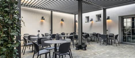 An outdoor patio area with round tables and chairs, a pergola structure, and wall-mounted lighting.