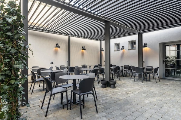 An outdoor patio area with round tables and chairs, a pergola structure, and wall-mounted lighting.