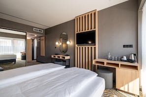 A hotel room with a large bed, a desk with a mirror, a television, and a kitchenette area.