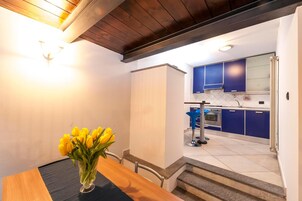 Un comedor con una mesa y un jarrón de flores amarillas, una cocina con armarios azules y una escalera que sube a un piso superior.