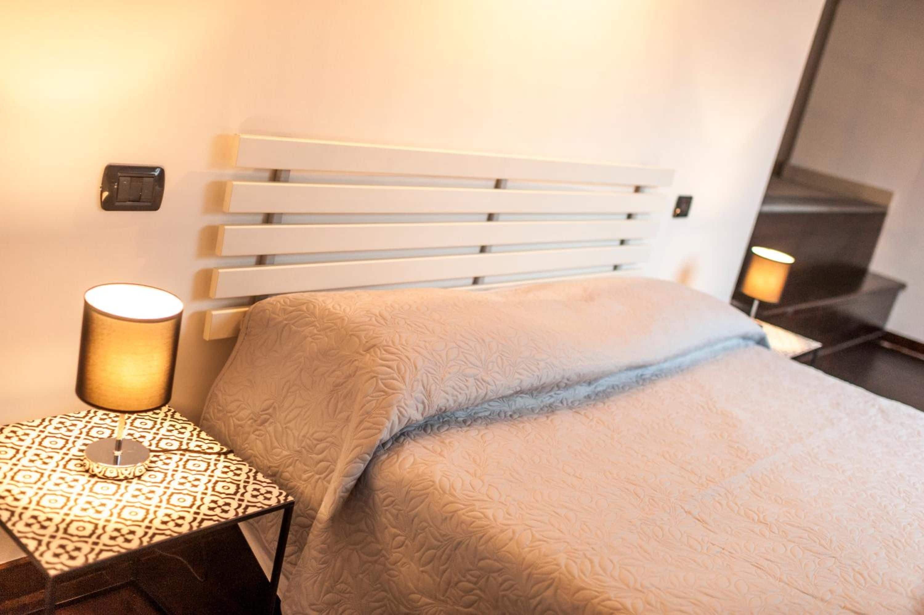 A hotel room with a bed, a bedside table, a lamp, and a wall-mounted switch.