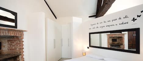 A bedroom with a brick fireplace, a bed with white bedding, a wall-mounted mirror, and a quote in Italian.