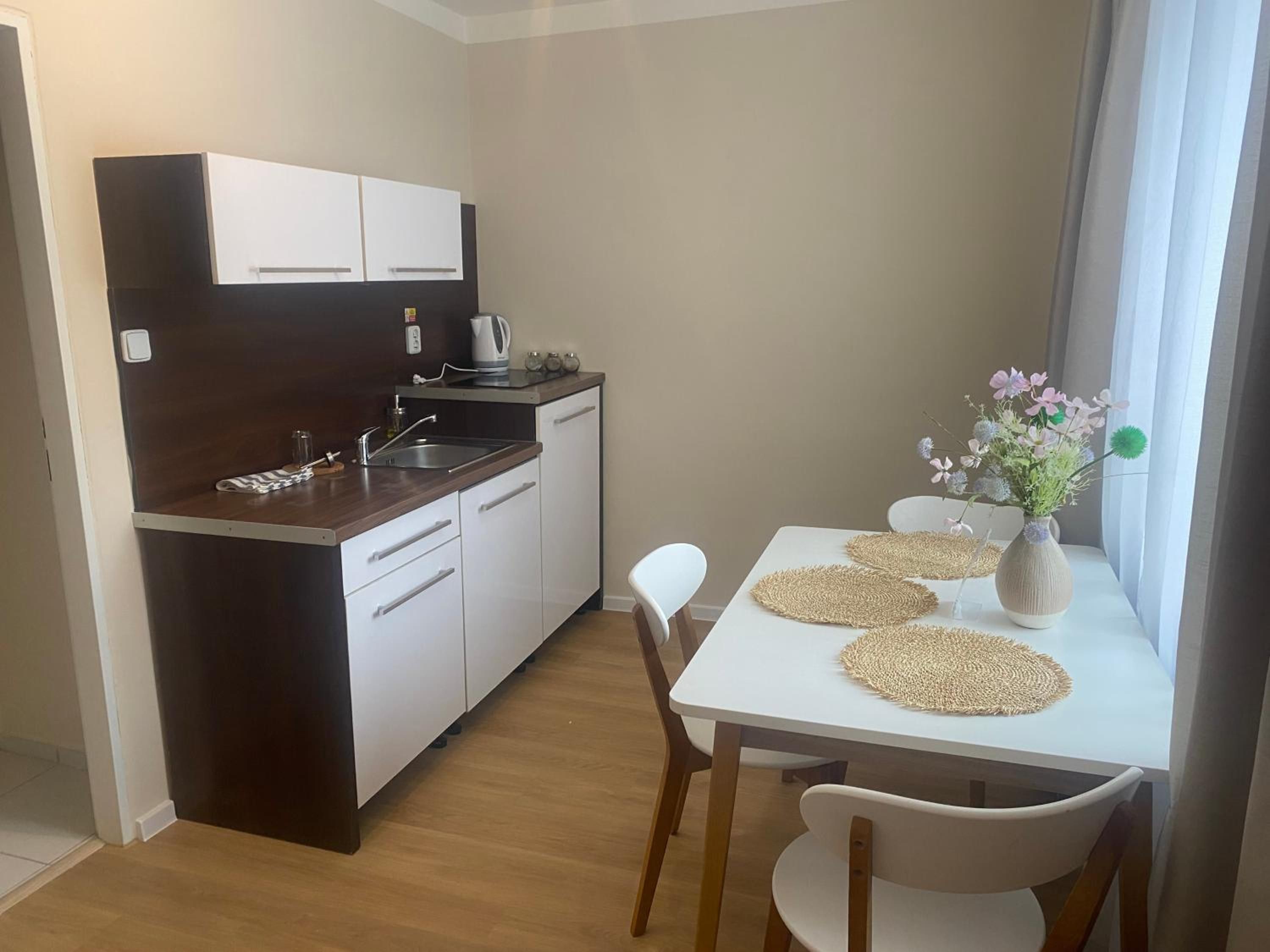 Una cocina compacta con armarios blancos, cafetera y una mesa de comedor con individuales y un florero.