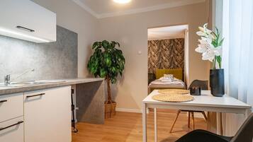 A modern kitchen with a built-in oven, a dining area with a table and chairs, a potted plant, and a view into a bedroom with a patterned wallpaper.