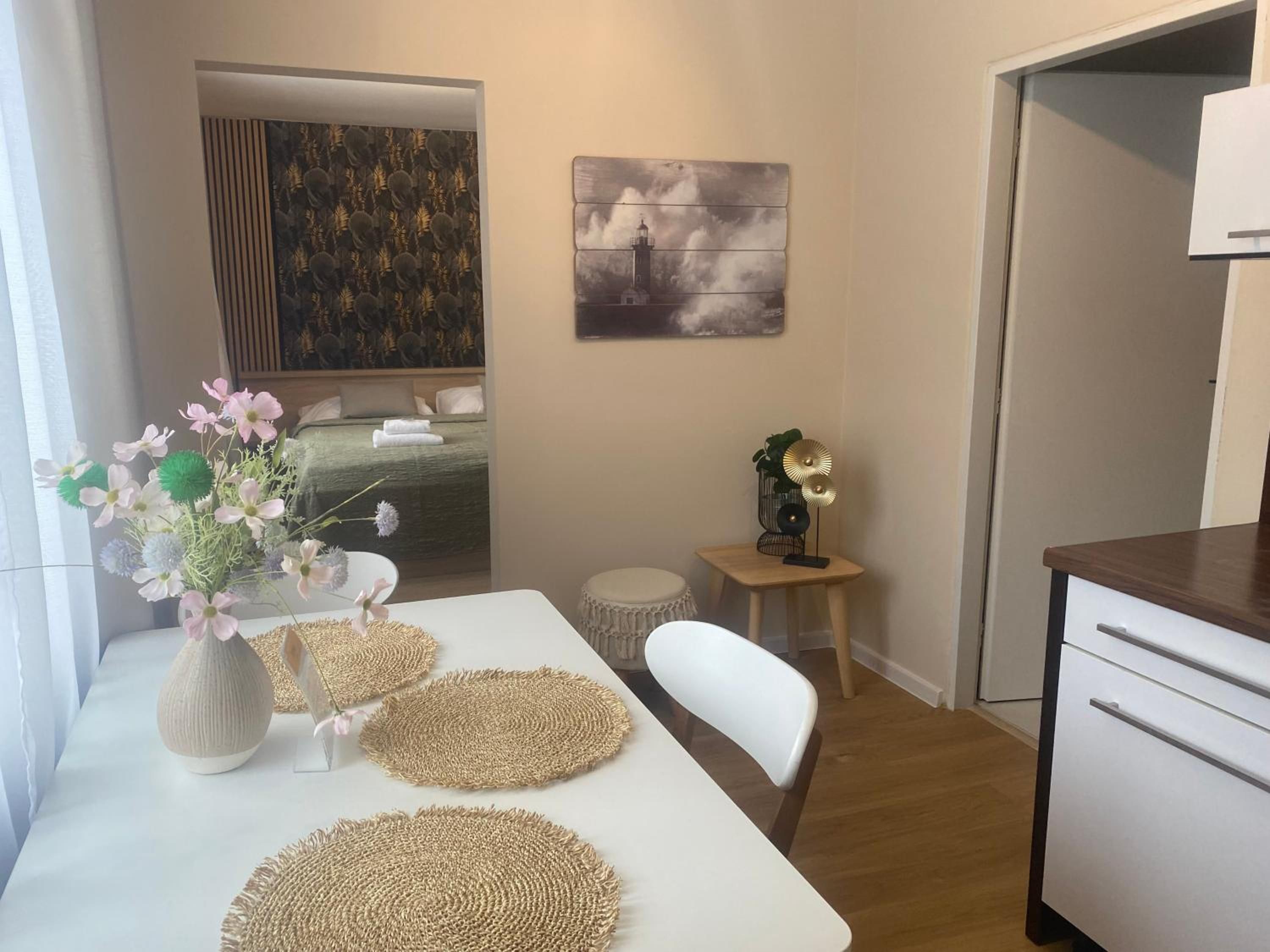 Una habitación pequeña y moderna con una mesa blanca, una silla, un taburete, un armario blanco y una cama con cabecera verde.
