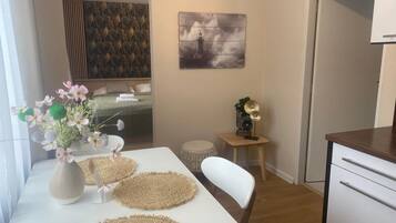Una habitación pequeña y moderna con una mesa blanca, una silla, un taburete, un armario blanco y una cama con cabecera verde.