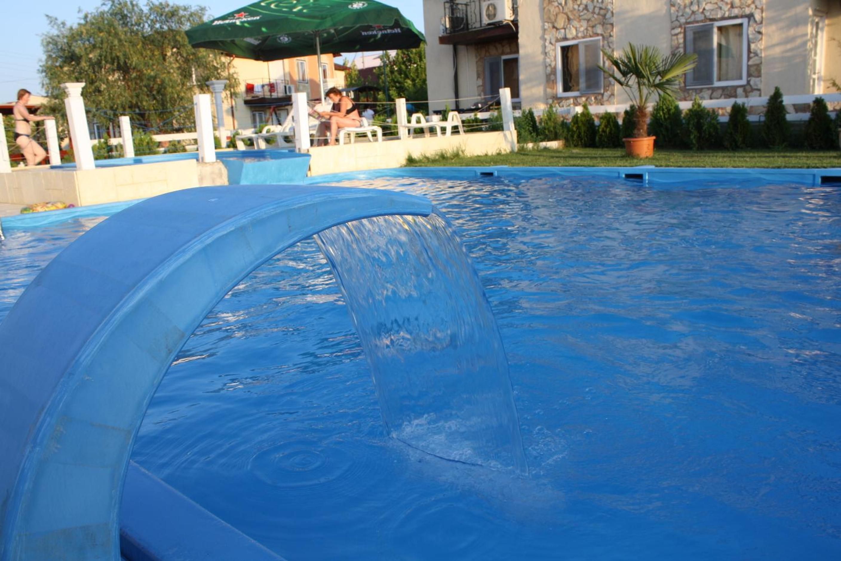 A swimming pool with a water feature, surrounded by a fence, with buildings and people in the background.