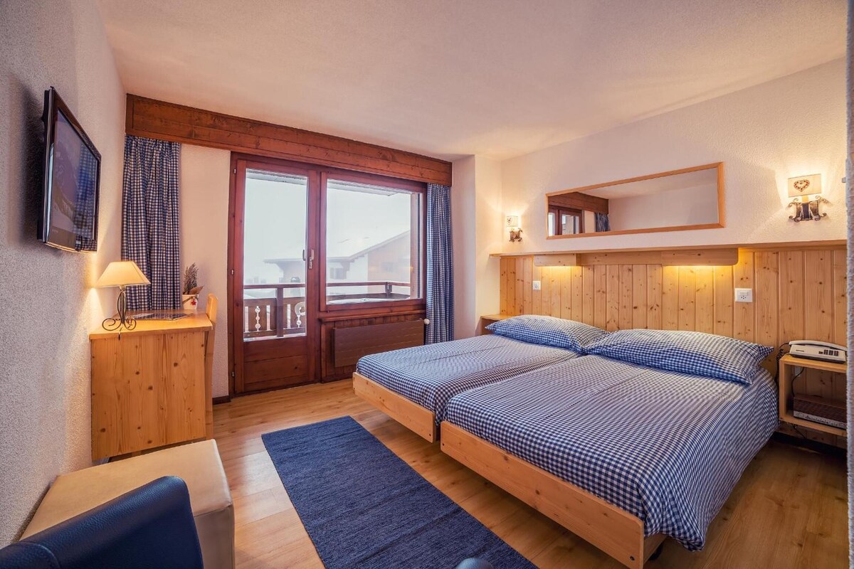 A bedroom with a bed, a desk, a chair, a TV, and a window with curtains.
