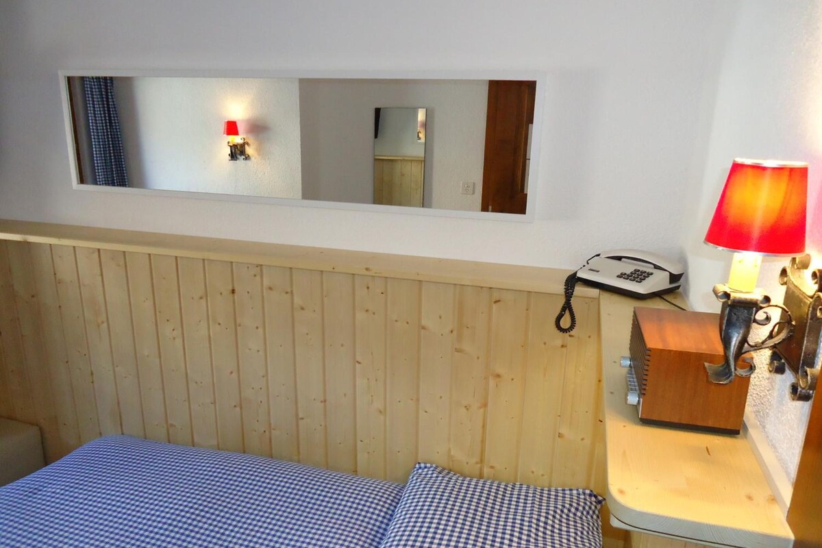 A room with a bed, a bedside table with a lamp, a telephone, and a mirror on the wall.